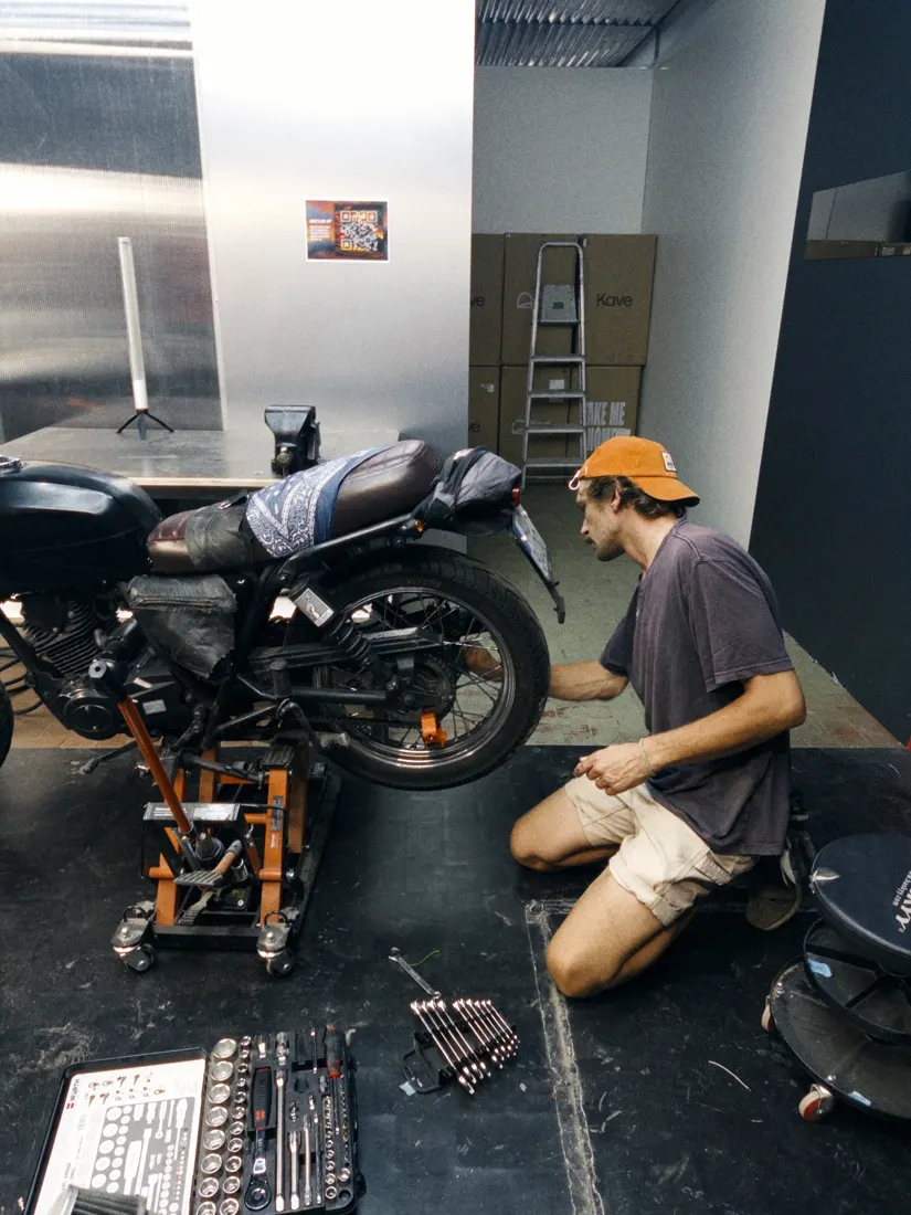 A mechanic in an orange cap kneeling by a bike on the lift, a tool case open on the floor